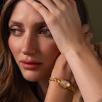 A woman with light brown hair rests her head on her hand, wearing the Chevron Diamond Link Timepiece in 18K Gold, looking thoughtful.
