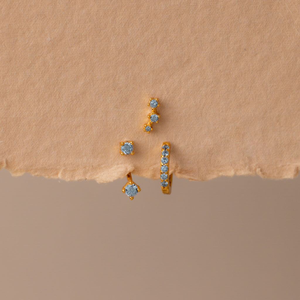 The Aquamarine Earring Trio Set features three gold earrings with blue gemstones—a delicate huggie hoop, an elegant ear climber, and a third design—displayed on textured beige paper.
