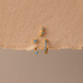 The Aquamarine Earring Trio Set features three gold earrings with blue gemstones—a delicate huggie hoop, an elegant ear climber, and a third design—displayed on textured beige paper.