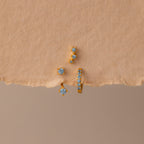 The Aquamarine Earring Trio Set features three gold earrings with blue gemstones—a delicate huggie hoop, an elegant ear climber, and a third design—displayed on textured beige paper.