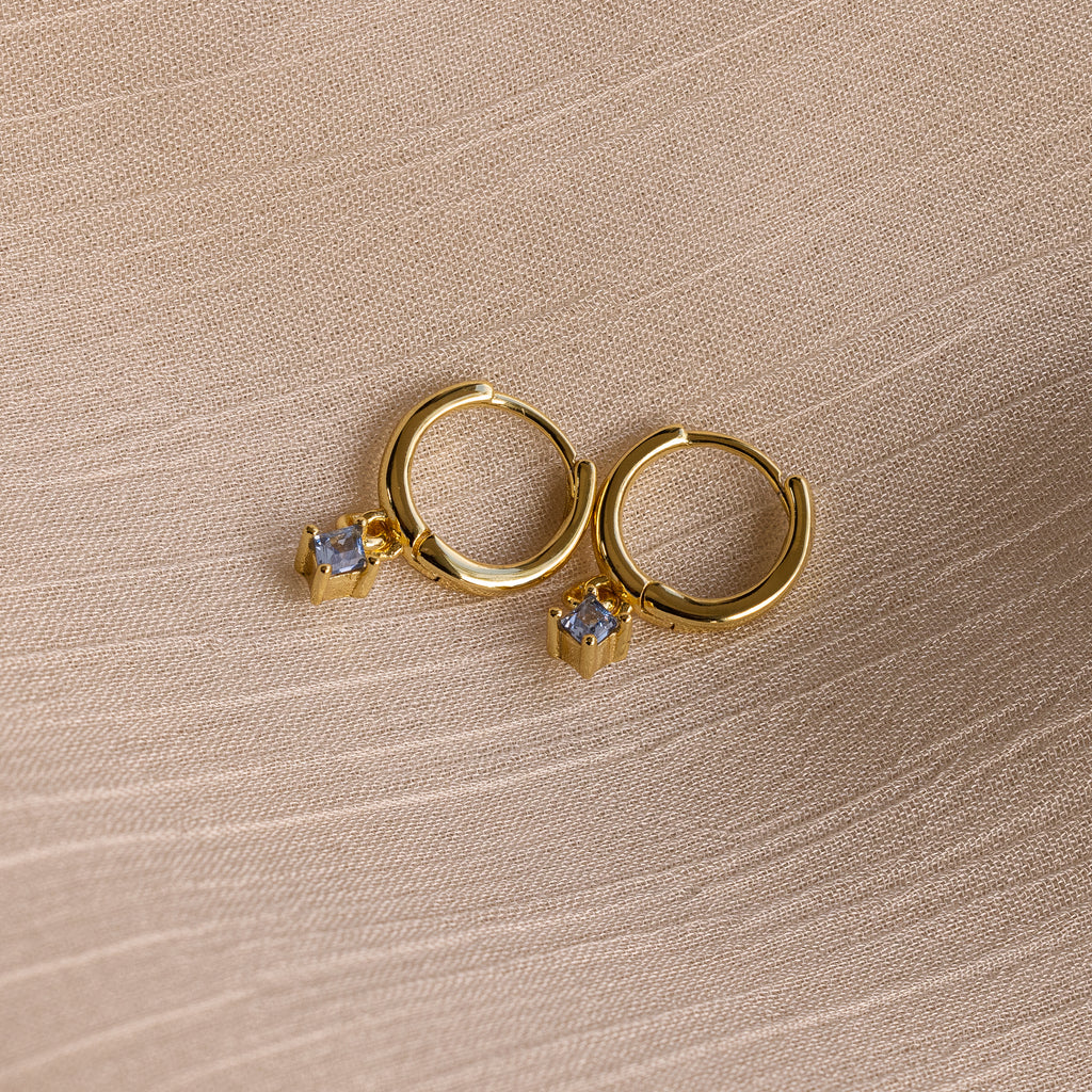 Birthstone Drop Huggies—gold hoop earrings with small square gemstone charms—make a stunning addition to your birthstone earring collection. Pictured on a textured beige fabric background.
