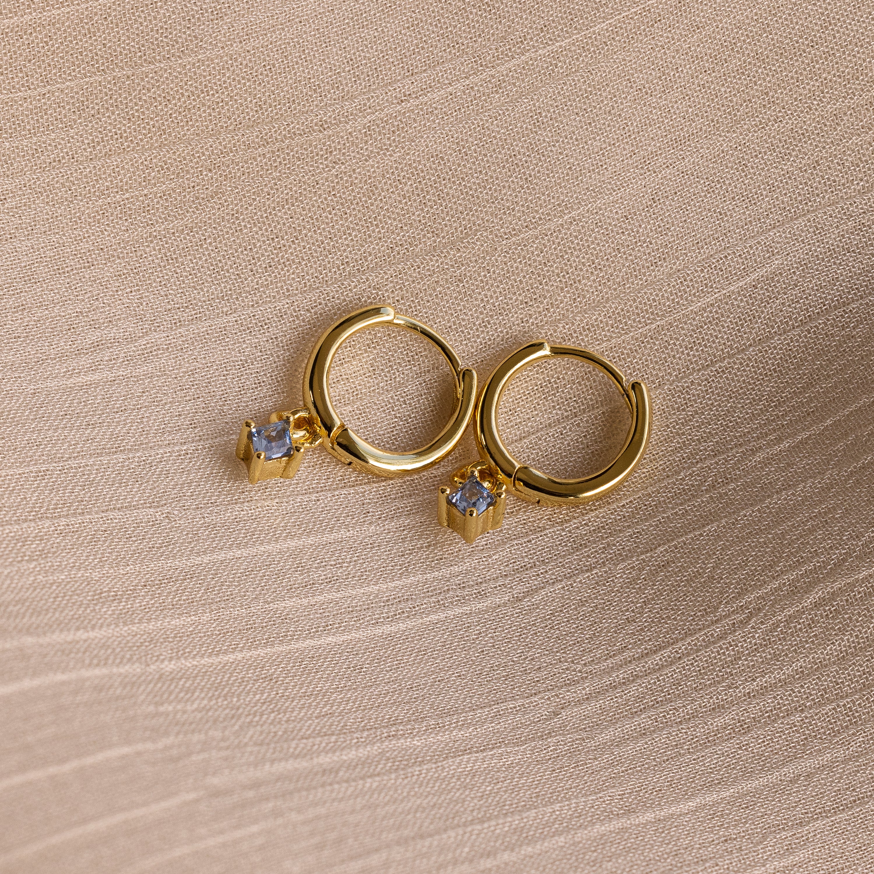 Birthstone Drop Huggies—gold hoop earrings with small square gemstone charms—make a stunning addition to your birthstone earring collection. Pictured on a textured beige fabric background.