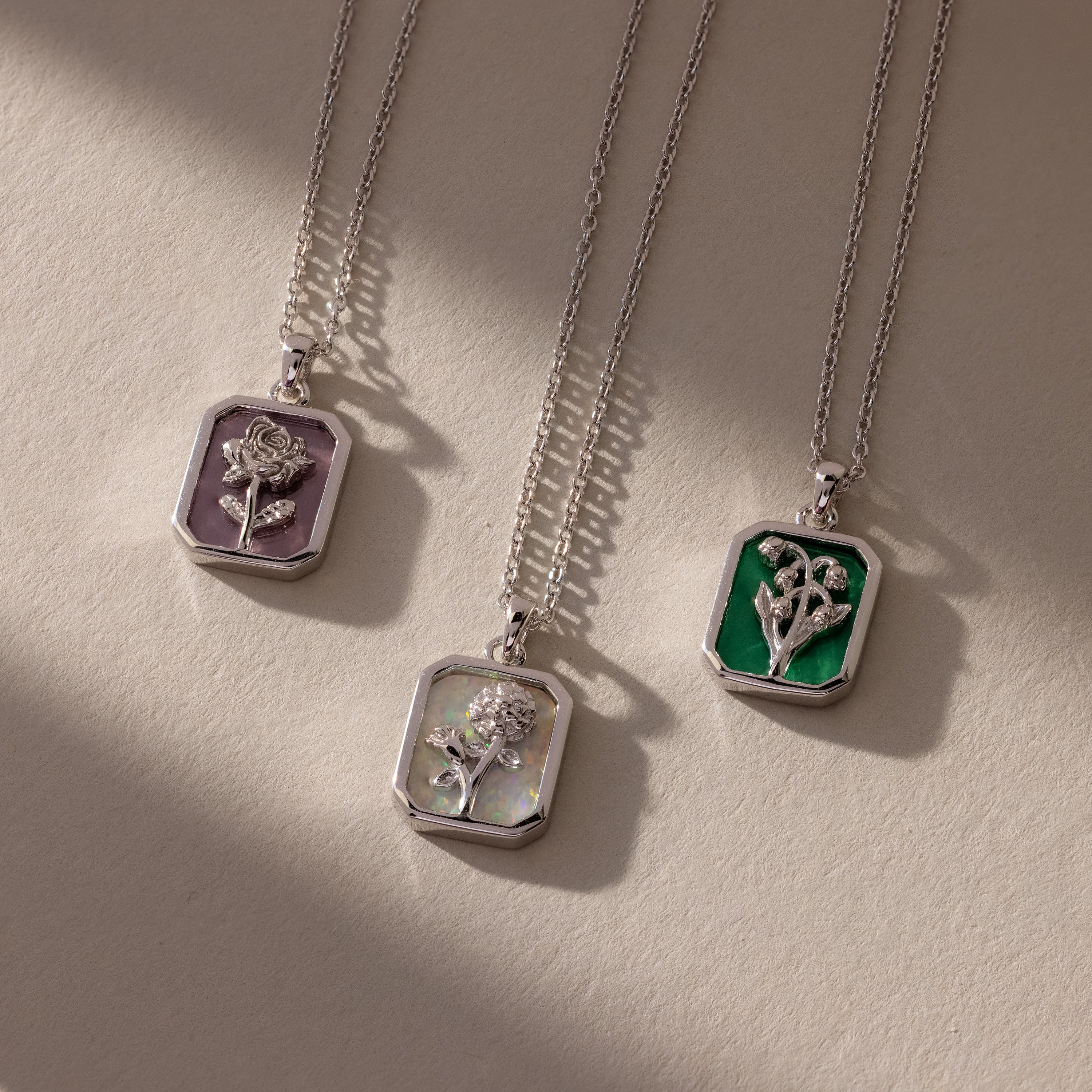Three Birth Flower Charm Necklaces shine on purple, iridescent, and green backgrounds—unique floral jewelry pieces that add a touch of elegance to any outfit.