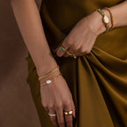 A woman in an olive dress wears gold and green jewelry, including rings, a luxury bracelet, the V-Link Chain Tennis Bracelet, and a watch.