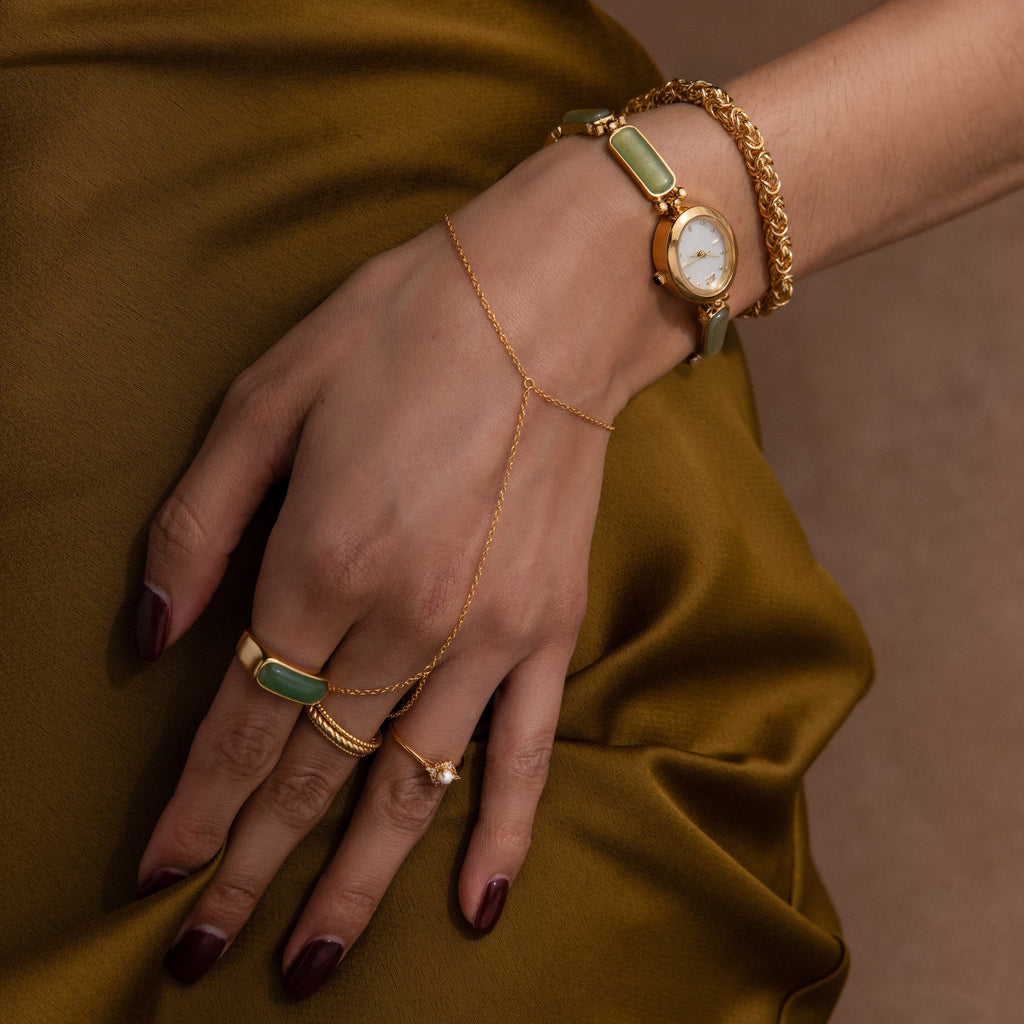 A hand wearing the Jade Band Timepiece with gold jewelry and green stones rests on olive satin fabric.