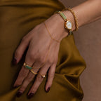 A hand wearing the Jade Band Timepiece with gold jewelry and green stones rests on olive satin fabric.