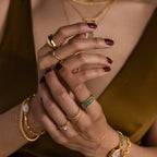 Woman with maroon nails, gold jewelry, and a Thin Stacking Rings Set with green stone and delicate bands gently clasping hands; olive top visible.