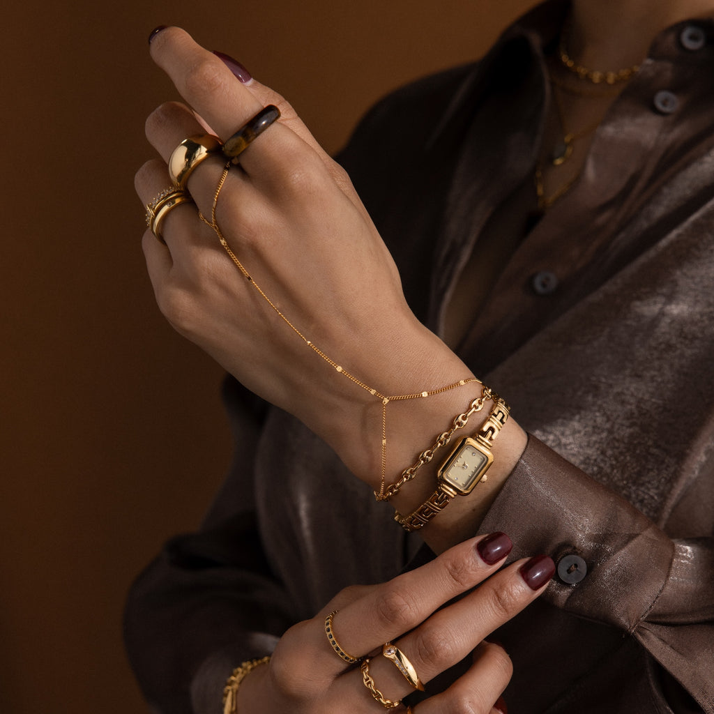 A close-up of hands with a Beaded Hand Chain Bracelet, gold bracelets, a watch, chain ring, dark nail polish, and a shiny brown shirt.