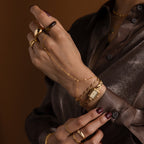 A close-up of hands with a Beaded Hand Chain Bracelet, gold bracelets, a watch, chain ring, dark nail polish, and a shiny brown shirt.