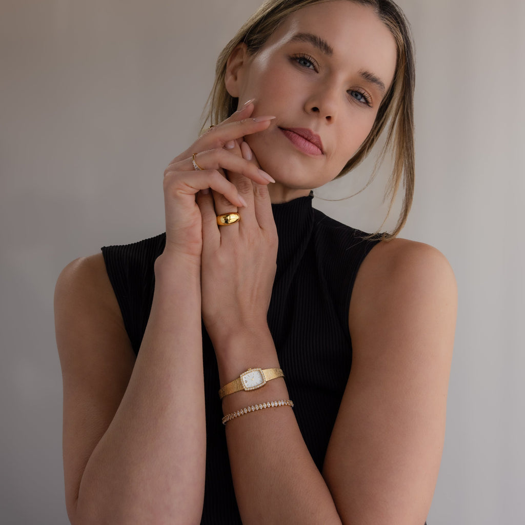 Woman in a sleeveless black top poses with her hands near her face, showcasing gold jewelry and the Petite Mesh Timepiece for a touch of modern elegance.