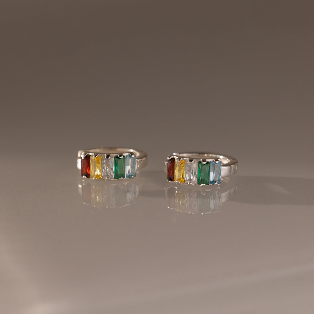 The Multiple Baguette Birthstone Huggies in Sterling Silver feature orange, yellow, clear, and green gemstones, creating a vibrant and colorful look beautifully displayed on a reflective surface.