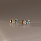 The Multiple Baguette Birthstone Huggies in Sterling Silver feature orange, yellow, clear, and green gemstones, creating a vibrant and colorful look beautifully displayed on a reflective surface.