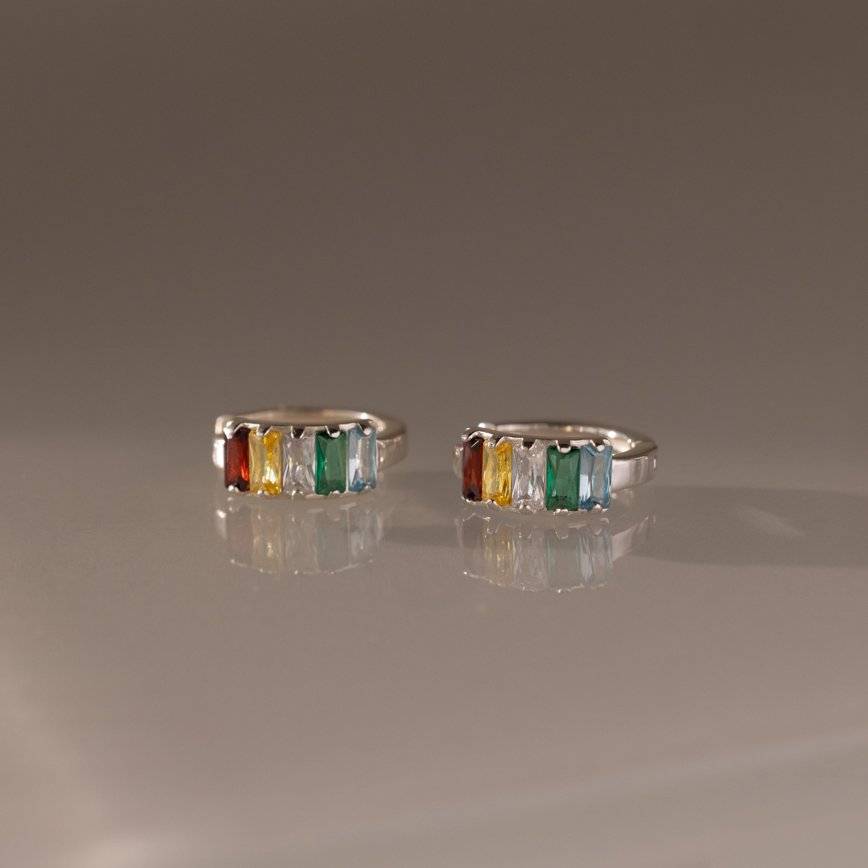 The Multiple Baguette Birthstone Huggies in Sterling Silver feature orange, yellow, clear, and green gemstones, creating a vibrant and colorful look beautifully displayed on a reflective surface.