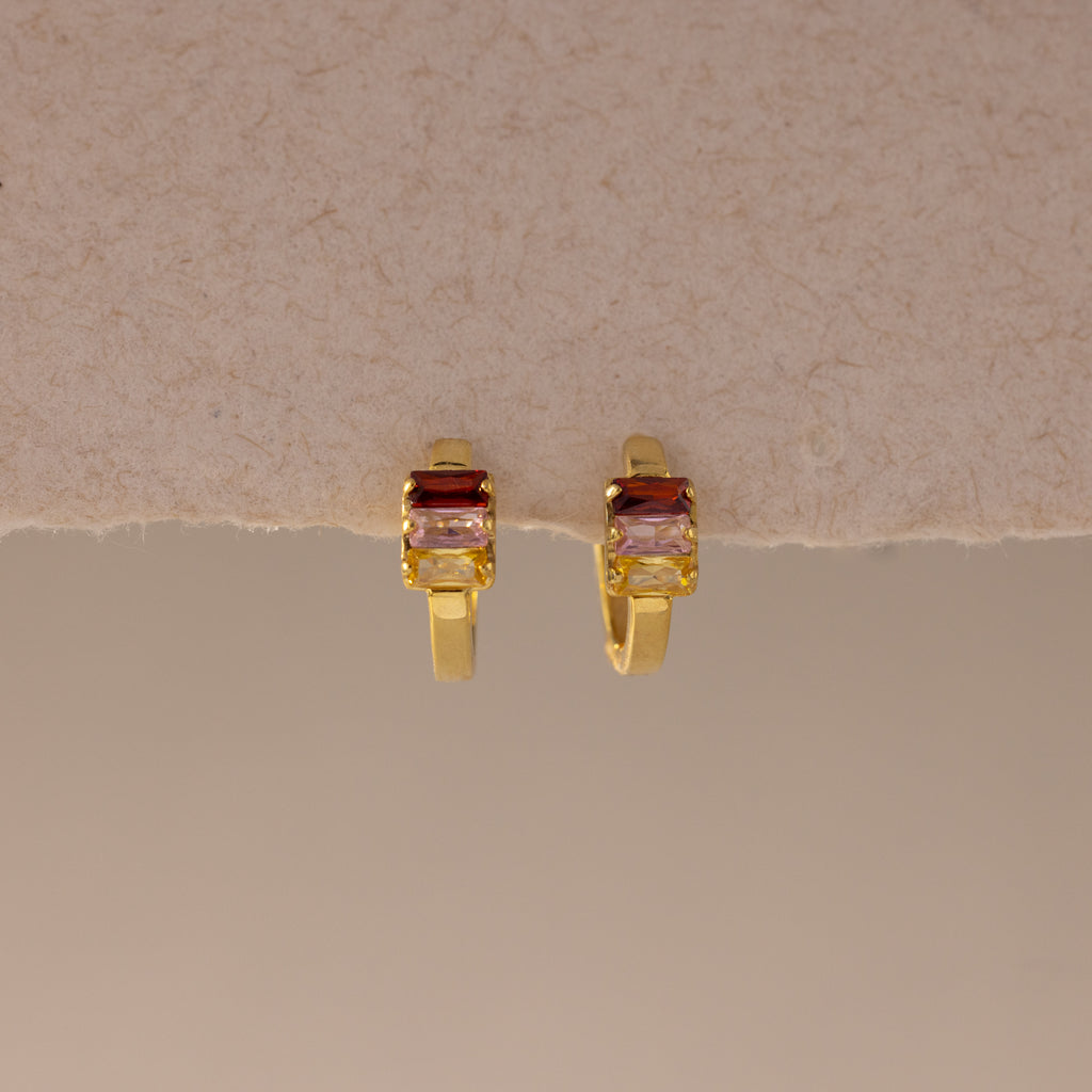 Multiple Baguette Birthstone Huggies in 18K Gold, featuring rectangular red and clear gemstones, are styled as colorful jewelry and displayed on a beige textured background.