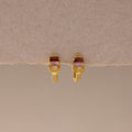 Multiple Baguette Birthstone Huggies in 18K Gold, featuring rectangular red and clear gemstones, are styled as colorful jewelry and displayed on a beige textured background.