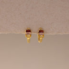 Multiple Baguette Birthstone Huggies in 18K Gold, featuring rectangular red and clear gemstones, are styled as colorful jewelry and displayed on a beige textured background.