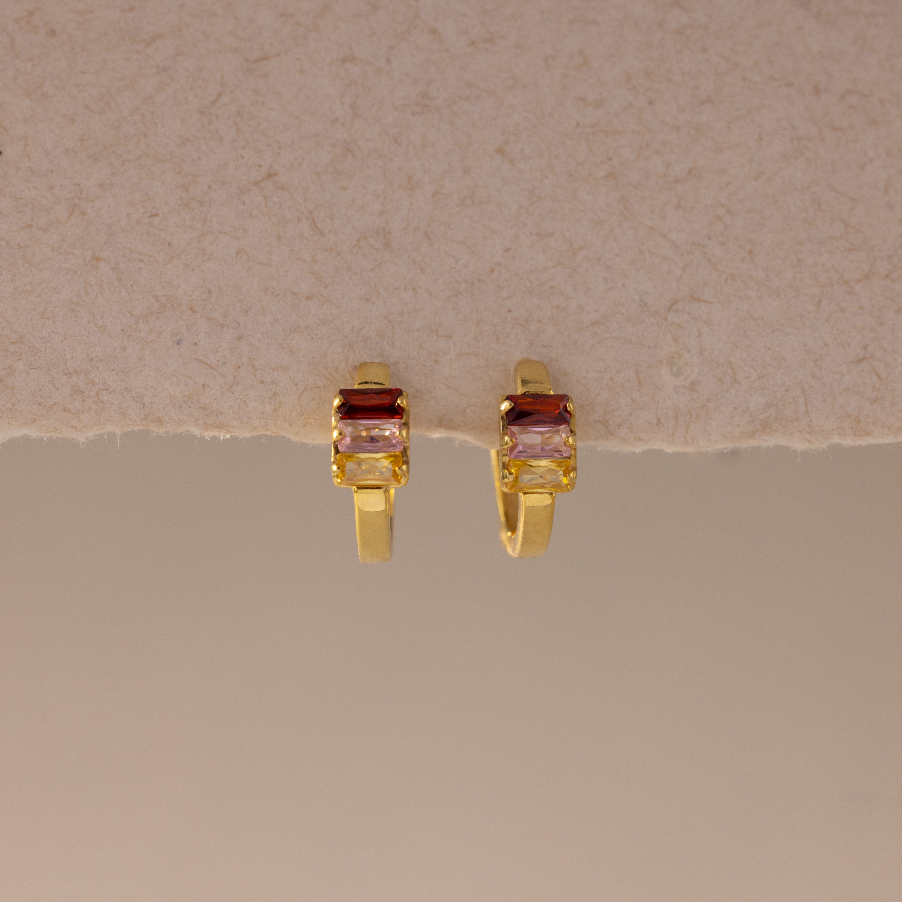 Multiple Baguette Birthstone Huggies in 18K Gold, featuring rectangular red and clear gemstones, are styled as colorful jewelry and displayed on a beige textured background.