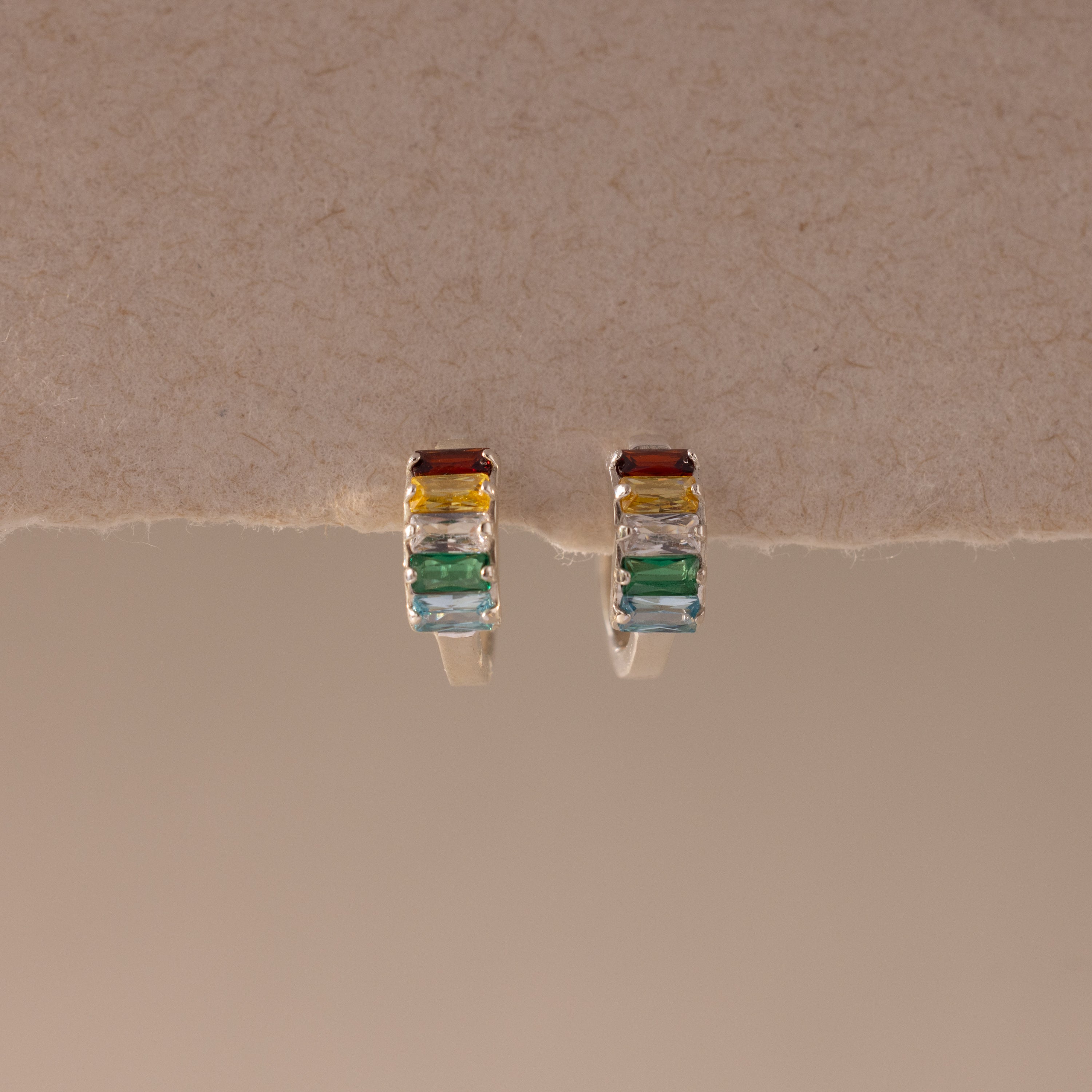 Multiple Baguette Birthstone Huggies in Sterling Silver, featuring rectangular multicolored gemstones, are beautifully showcased on a textured beige surface.