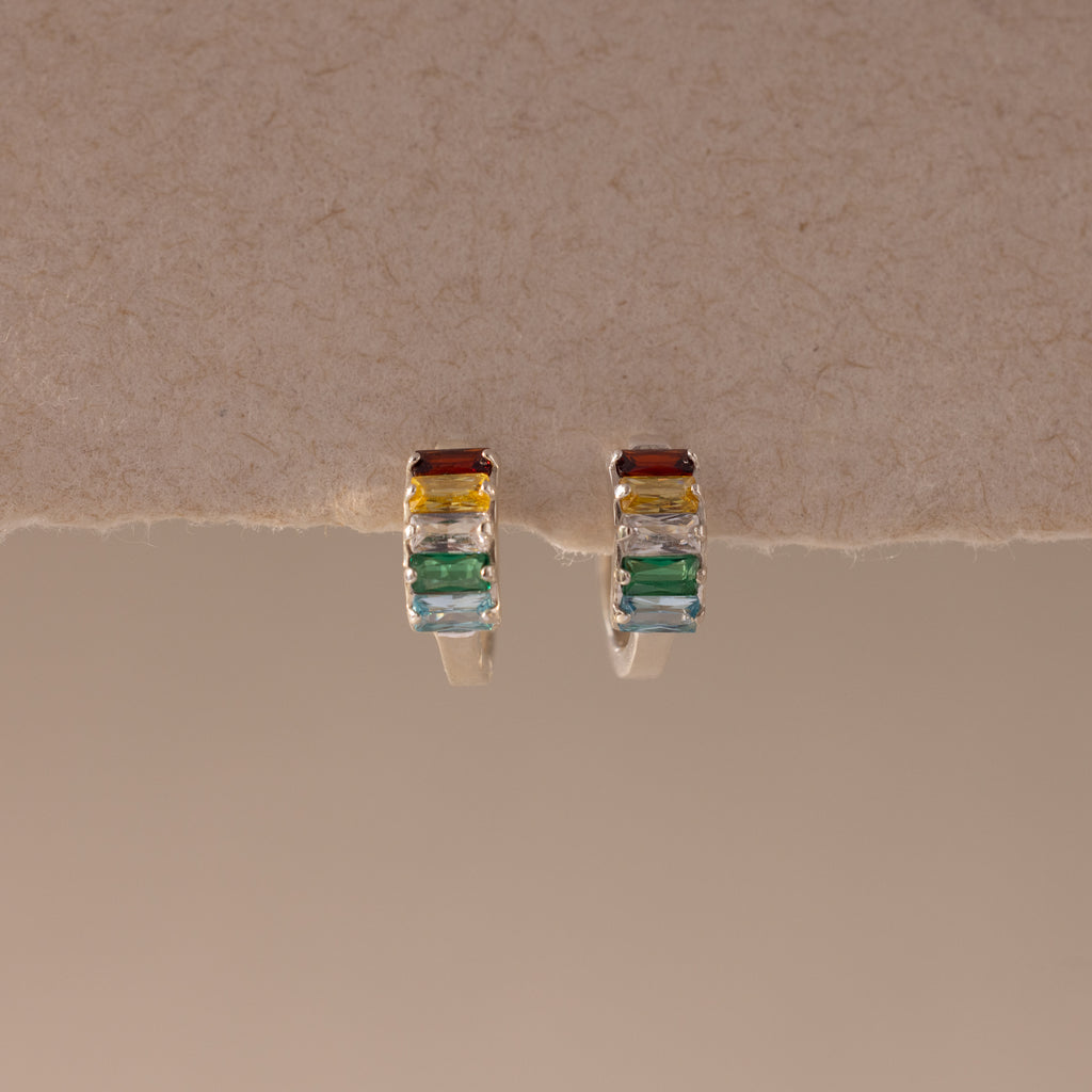 The Multiple Baguette Birthstone Huggies—silver petite hoops with rectangular red, yellow, blue, and green stones—rest on a textured beige surface, offering colorful jewelry for a playful accent to any look.