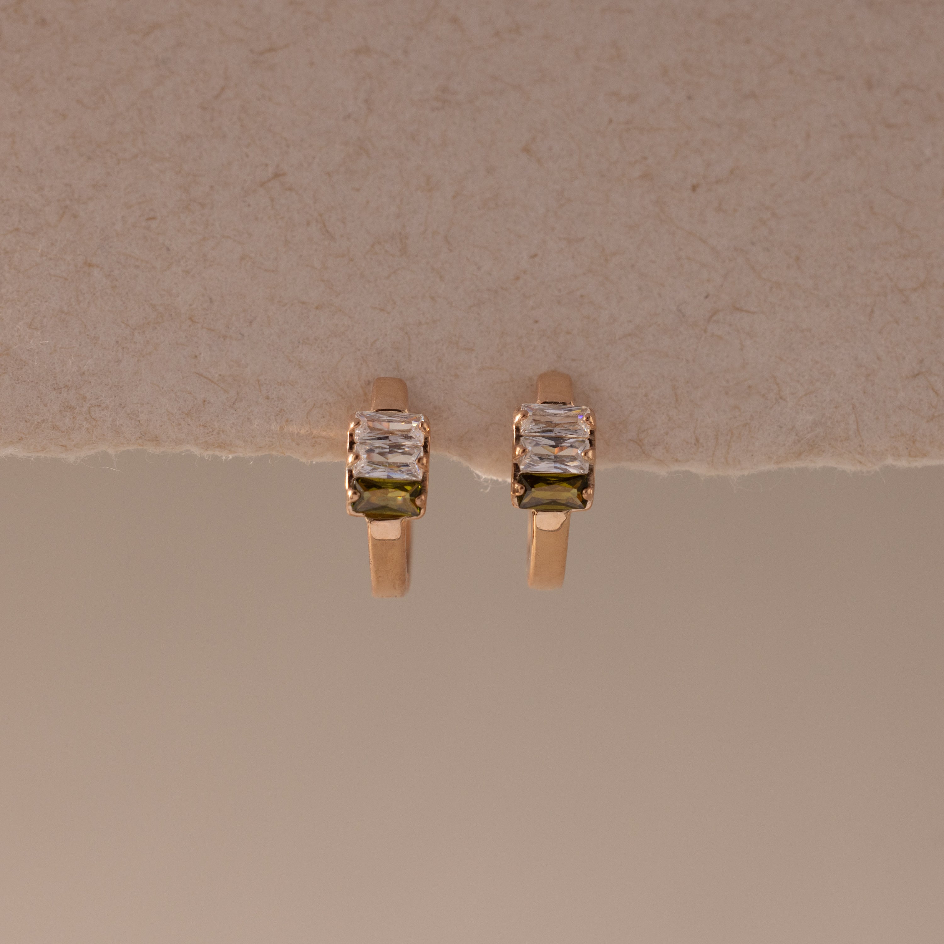 Multiple Baguette Birthstone Huggies in Rose Gold, featuring petite rectangular stones, are displayed on a textured beige background, combining timeless elegance with a modern feel.