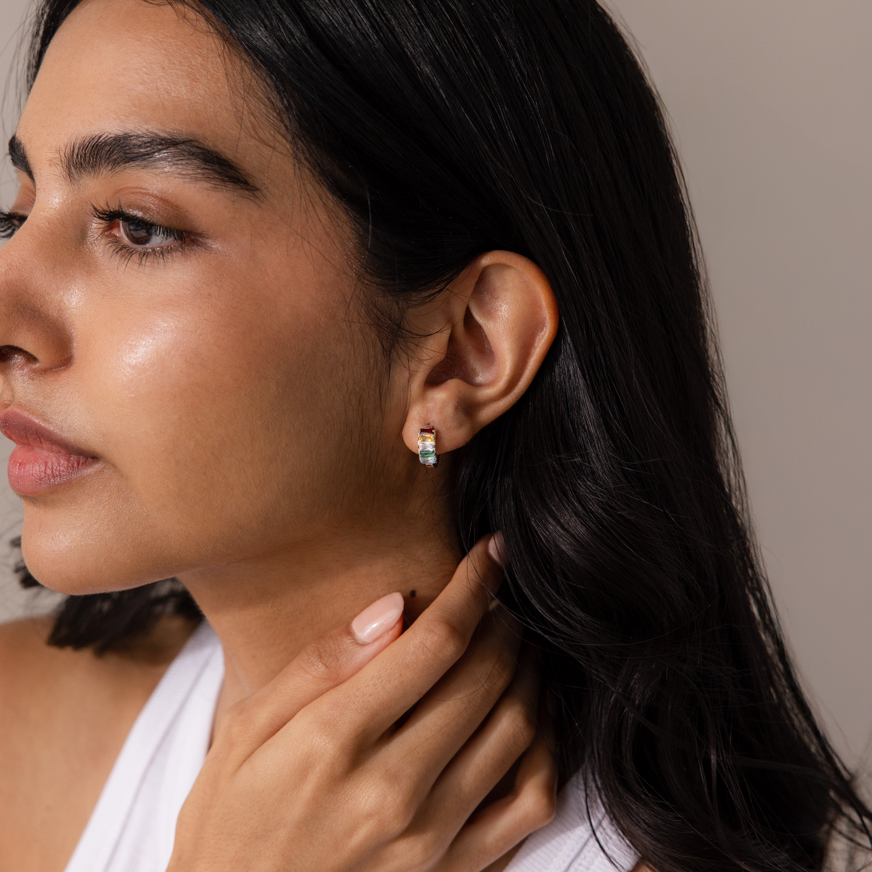 Woman with long dark hair wears Multiple Baguette Birthstone Huggies in Sterling Silver, touching her neck against a neutral background.