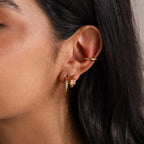 Close-up of a woman's ear with straight dark hair, wearing Multiple Baguette Birthstone Huggies in 18K Gold and a colorful stud.