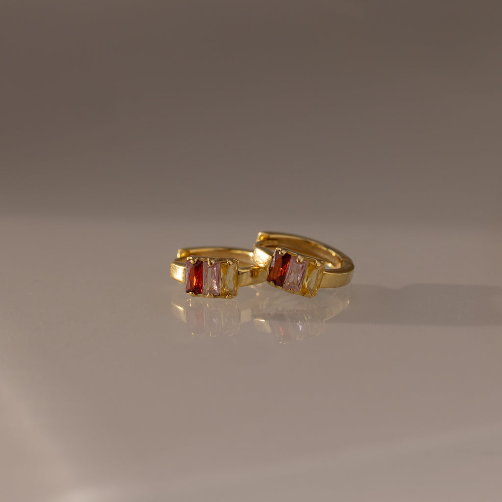The Multiple Baguette Birthstone Huggies in 18K Gold, featuring rectangular red and clear gemstones, are elegantly displayed on a smooth, reflective surface, highlighting their colorful and refined design.