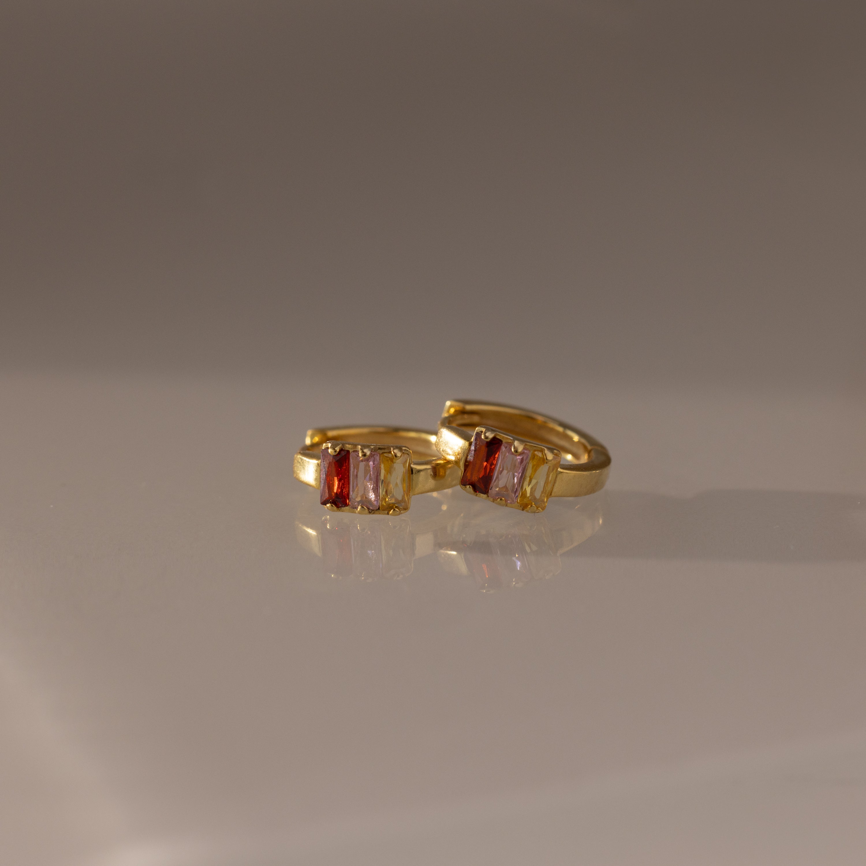 The Multiple Baguette Birthstone Huggies in 18K Gold, featuring rectangular red and clear gemstones, are elegantly displayed on a smooth, reflective surface, highlighting their colorful and refined design.