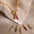 Gold chain necklace with heart and beaded charms, plus six colorful personalized pendants—including the Ombre Lace Birthstone Charm—displayed on a beige cloth.