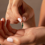 A person with manicured nails holds a silver earring with green gemstones, showcasing the elegant Ombre Lace Emerald Charm between their fingers.