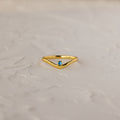 The Blue Zircon Drop Curve Ring, a gold stacking ring with a small blue gemstone, is displayed on a textured white surface.