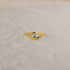 The Blue Zircon Drop Curve Ring, a gold stacking ring with a small blue gemstone, is displayed on a textured white surface.