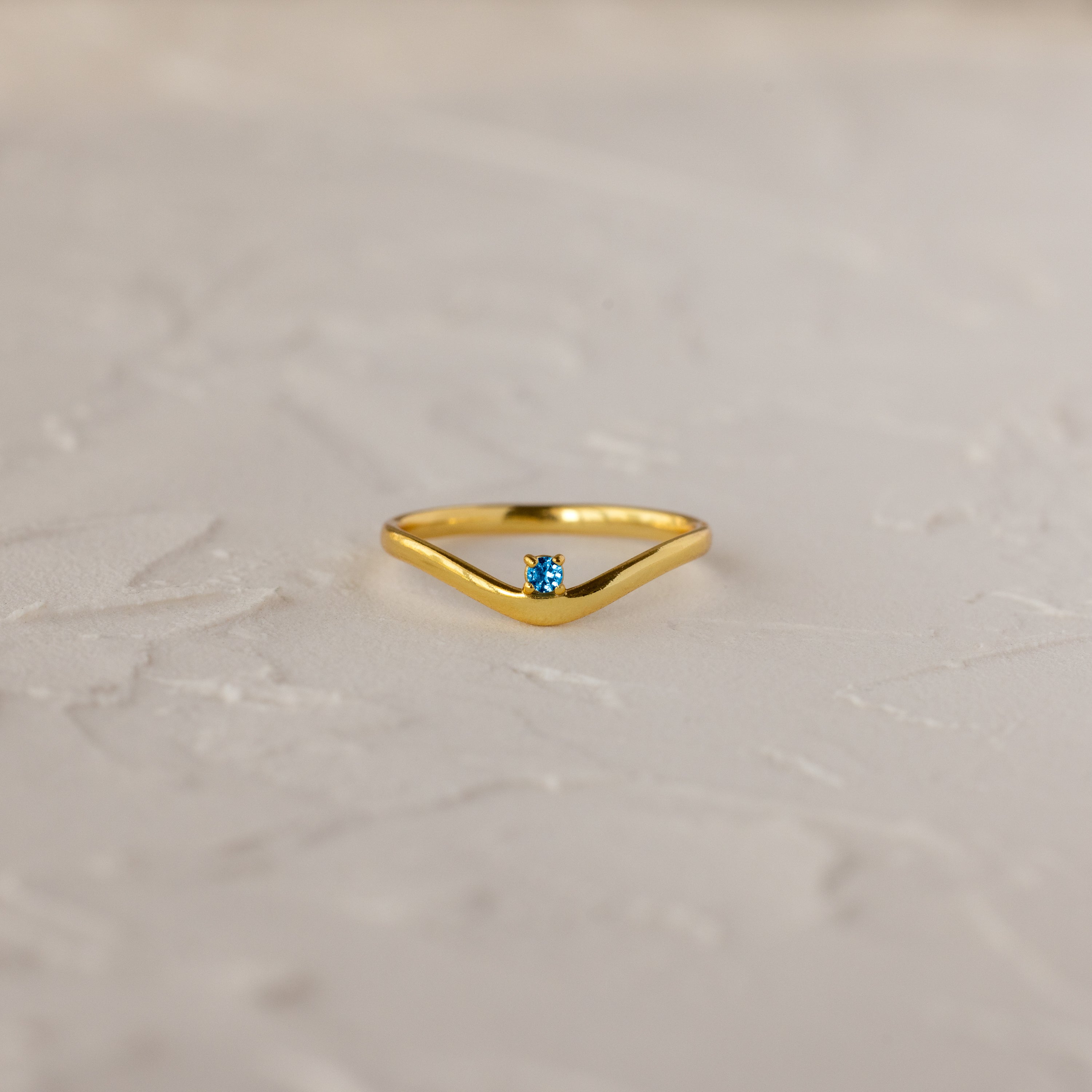 The Blue Zircon Drop Curve Ring, a gold stacking ring with a small blue gemstone, is displayed on a textured white surface.