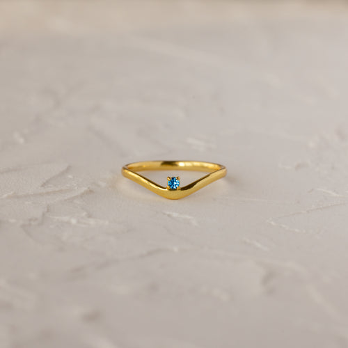 The Blue Zircon Drop Curve Ring, a gold stacking ring with a small blue gemstone, is displayed on a textured white surface.