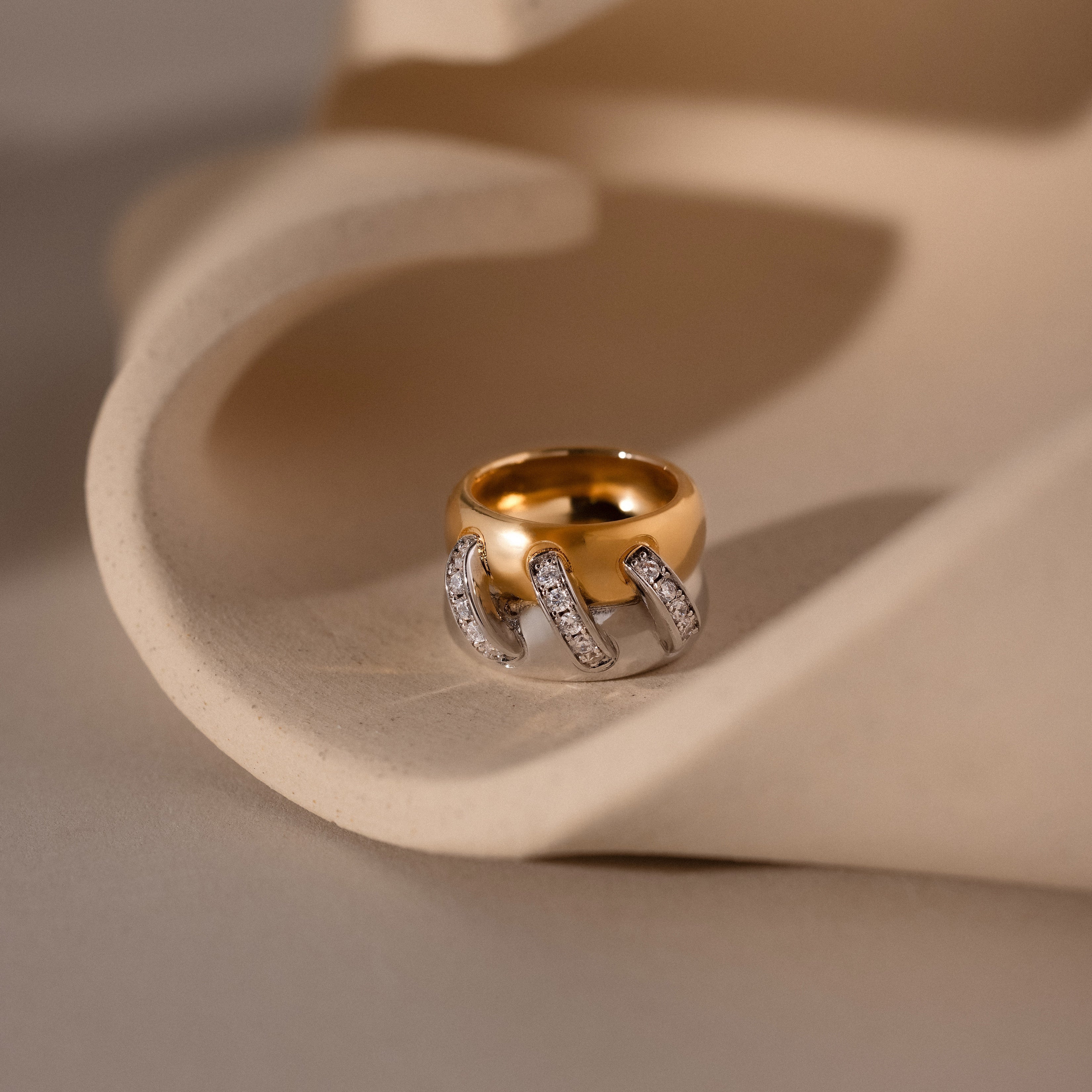 The Sculpted Mixed Metal Link Ring showcases gold and silver links accented with pavé diamonds, elegantly presented on a curved beige surface.