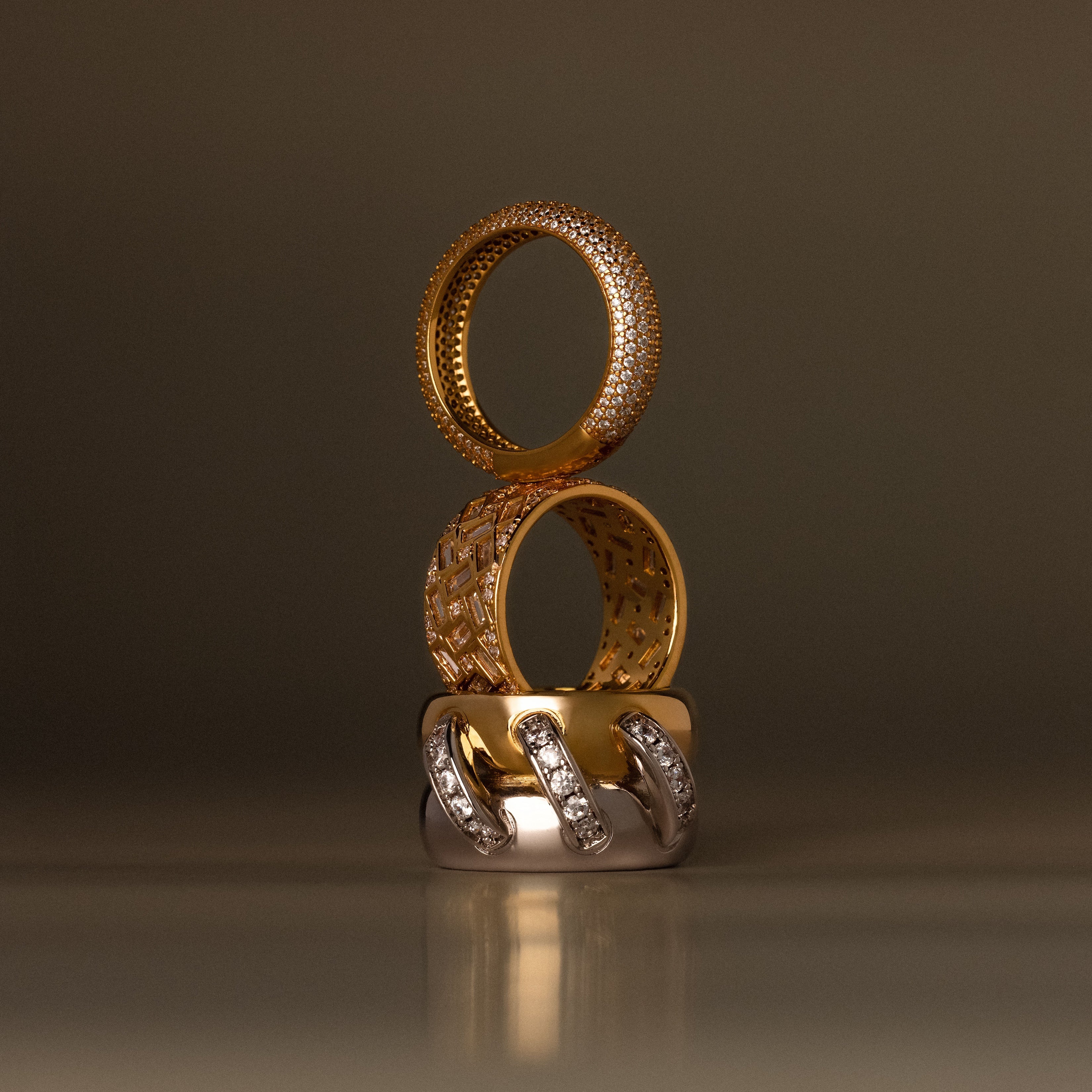 The Sculpted Mixed Metal Link Ring, adorned with pavé diamonds, is stacked with two other gold and diamond rings against a blurred brown background.