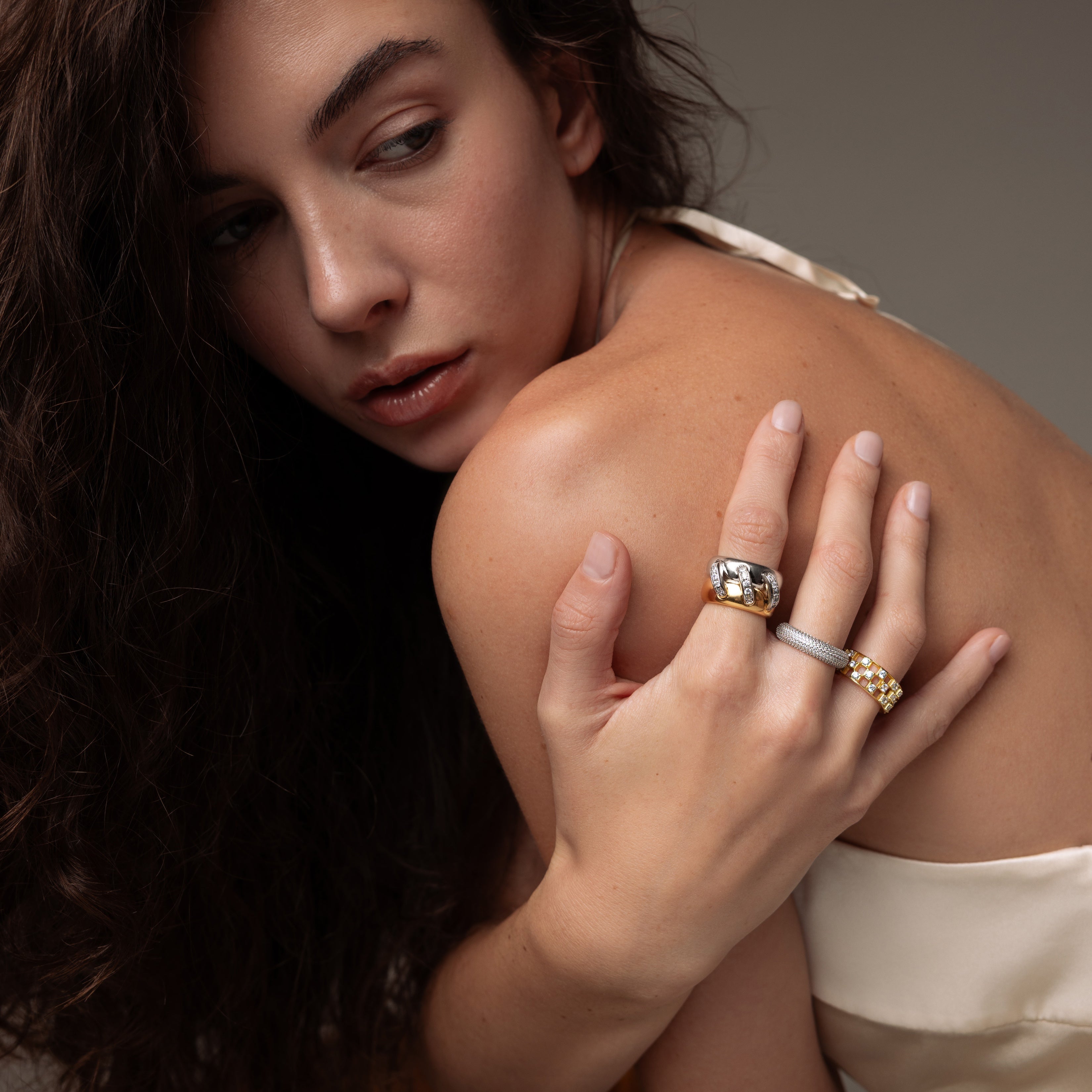 A woman with long dark hair glances over her shoulder, wearing a satin top and displaying the Sculpted Mixed Metal Link Ring adorned with pavé diamonds on her finger.
