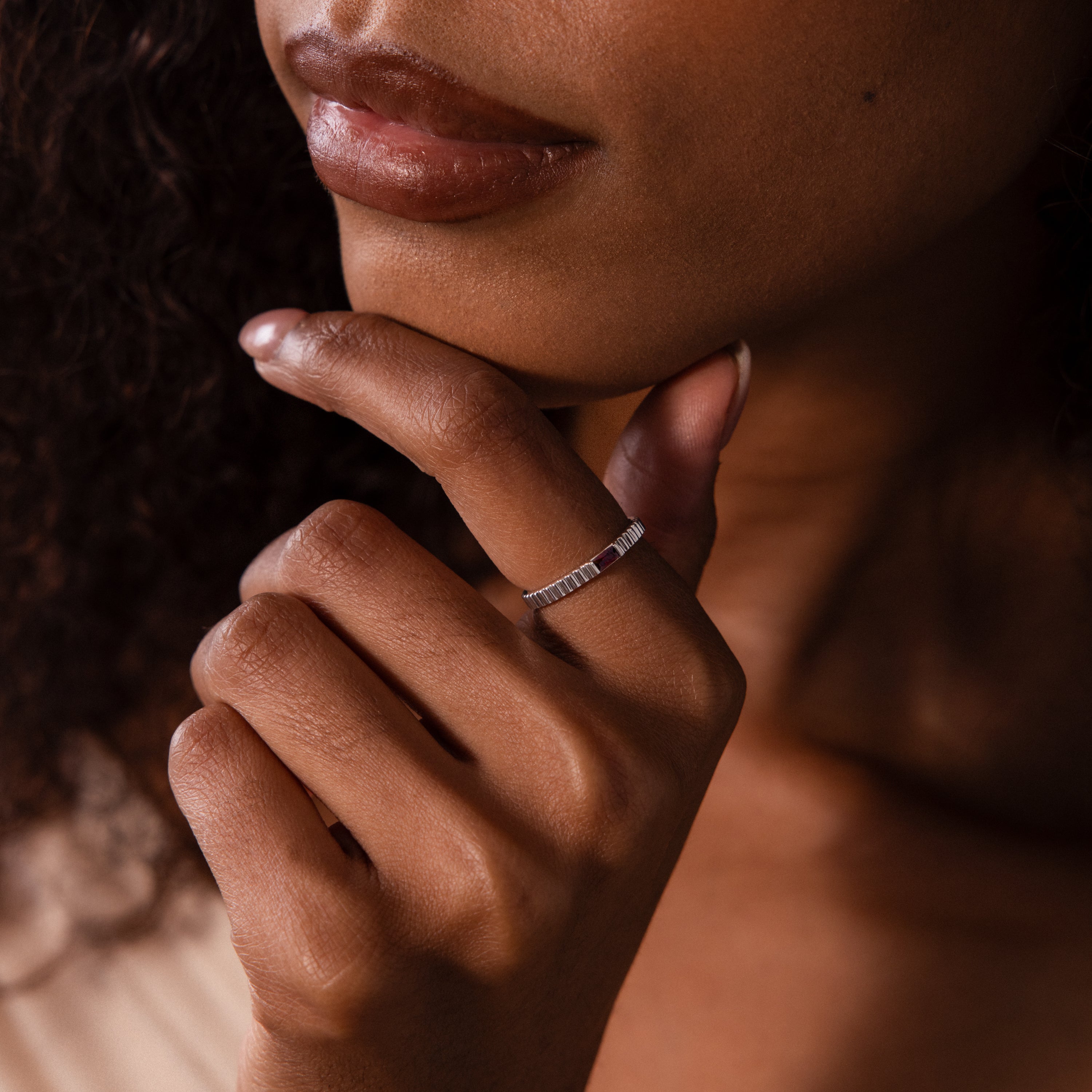 A woman with curly hair gently touches her chin, showcasing the Dainty Ribbed Ruby Ring on her finger—the delicate ruby adds a personal, vibrant touch to her look.