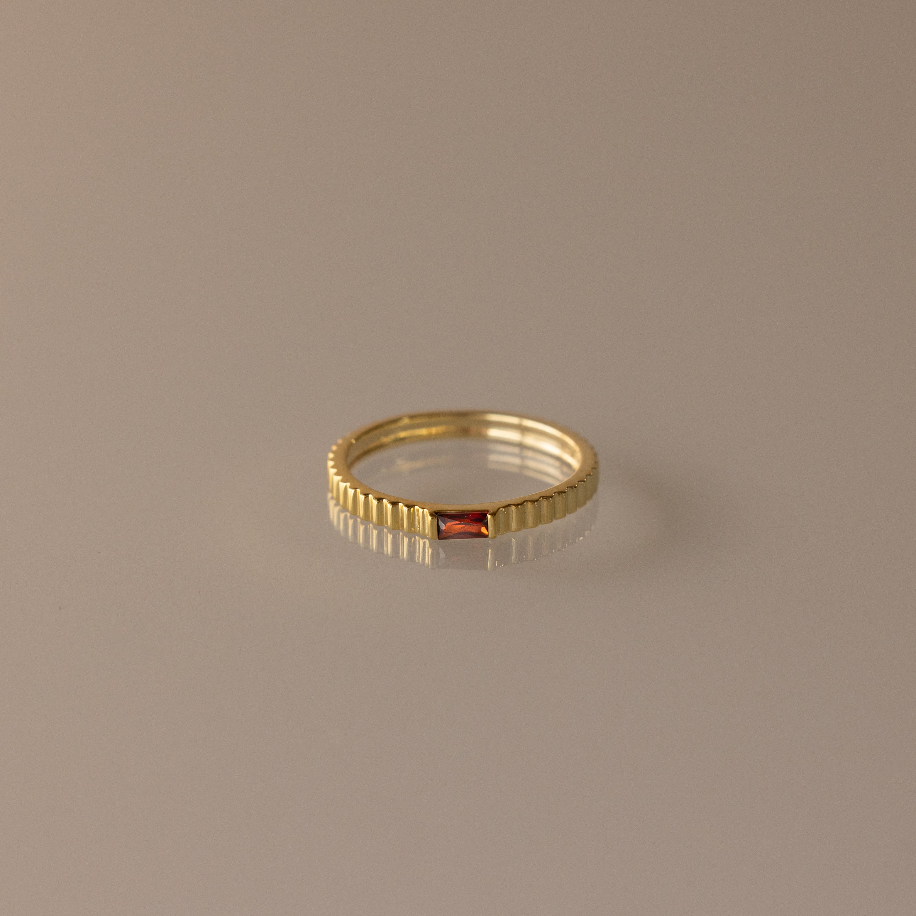 The Dainty Ribbed Garnet Ring features a ridged gold band with a small rectangular red gemstone, ideal as a delicate ring or personalized birthstone accessory, and is displayed on a neutral background.
