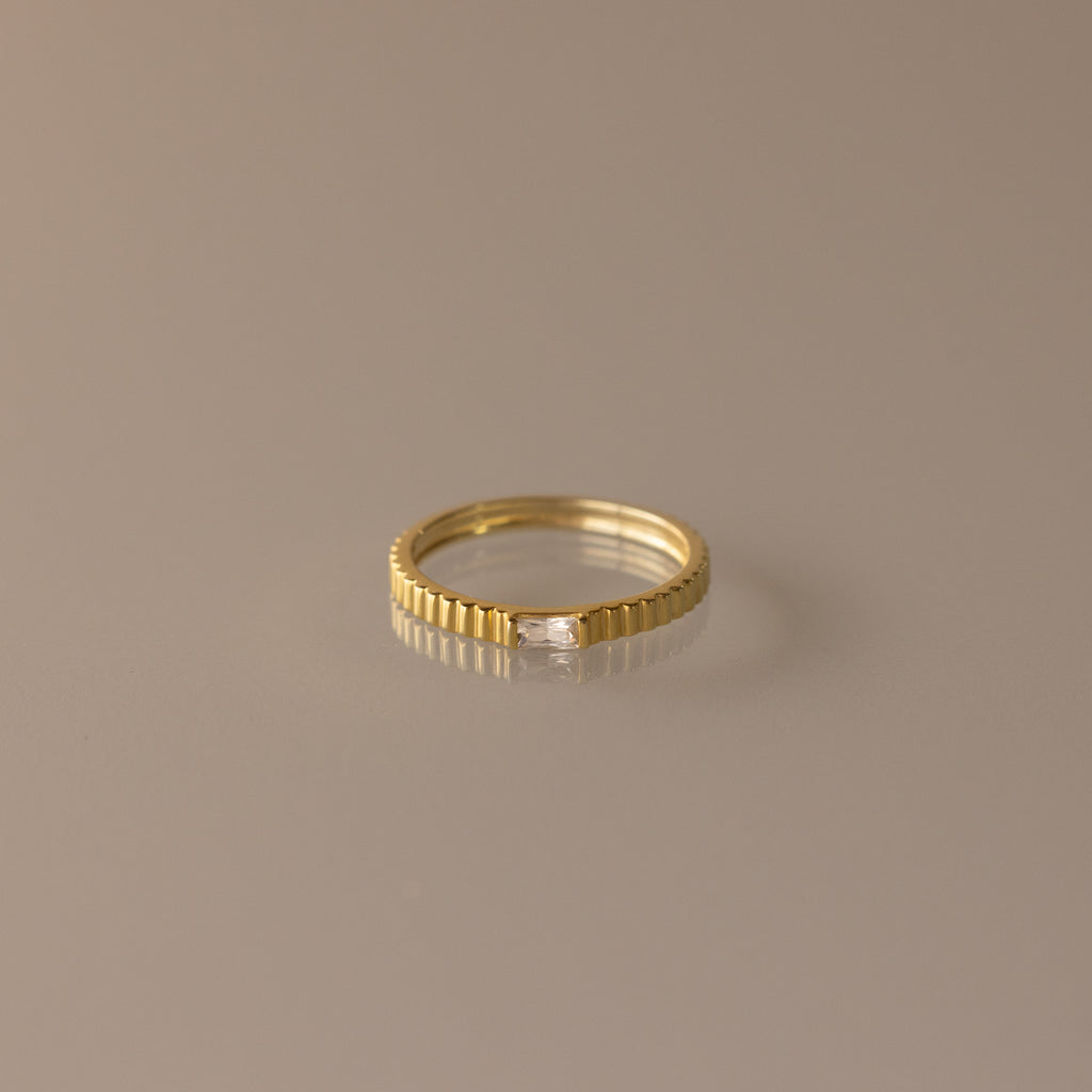 The Dainty Ribbed Diamond Ring features a slim gold ridged band and a petite rectangular diamond, perfect as a birthstone ring and shown on a neutral background.