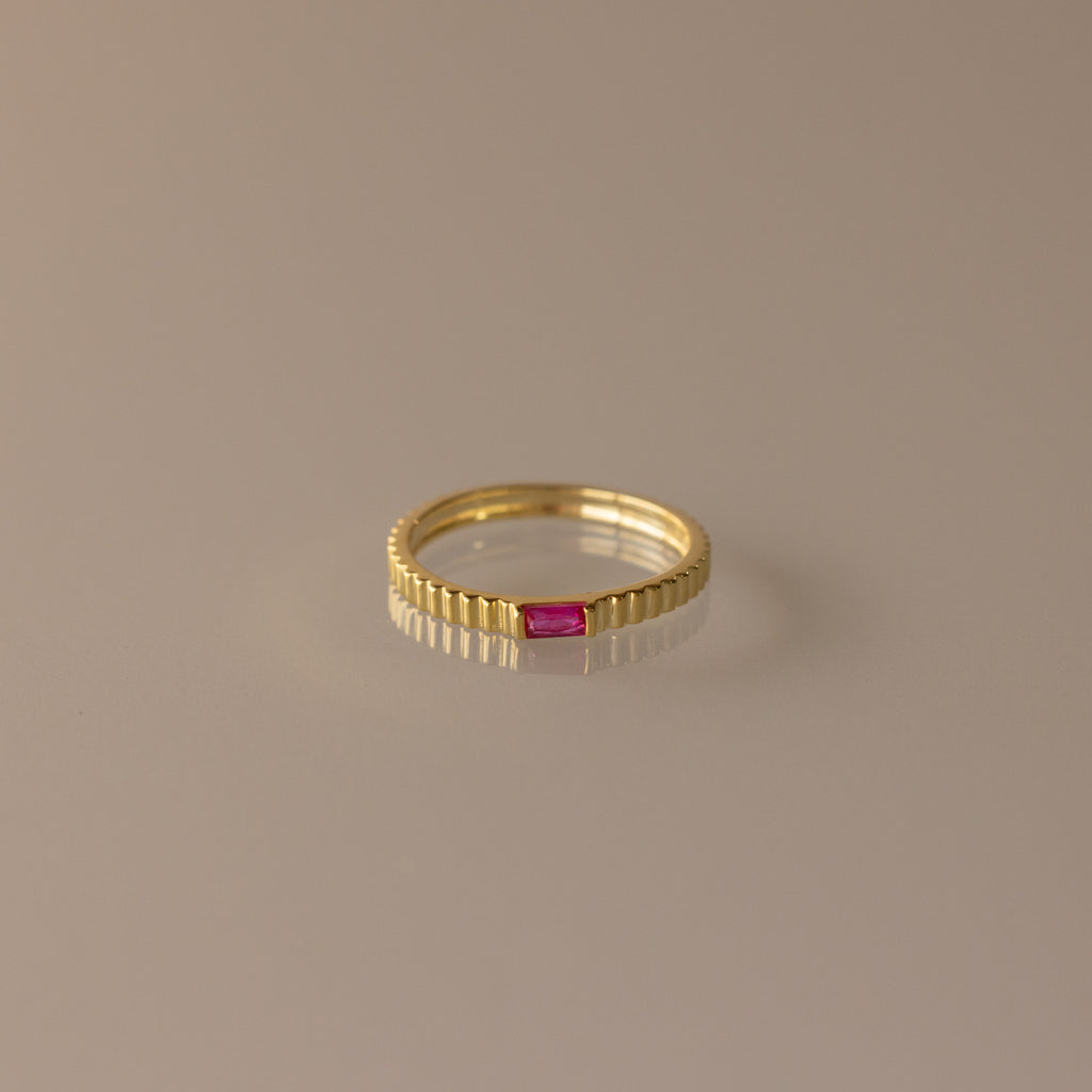 The Dainty Ribbed Ruby Ring features a gold ribbed band with a small rectangular pink gemstone at the center, ideal as a birthstone ring or for stacking.