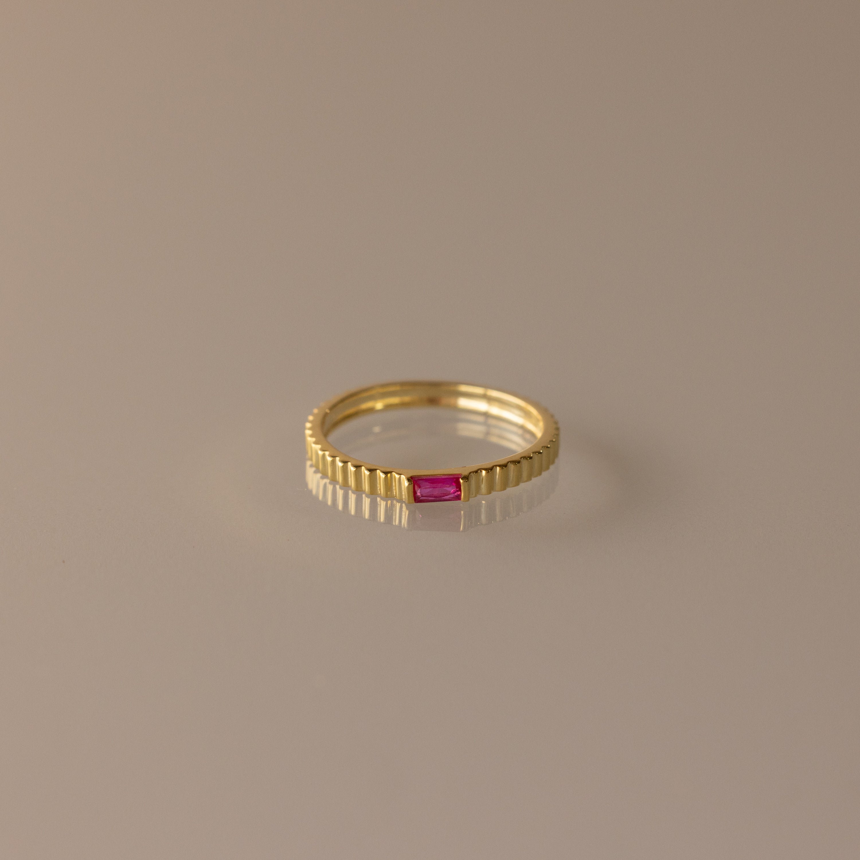 The Dainty Ribbed Ruby Ring features a gold ribbed band with a small rectangular pink gemstone at the center, ideal as a birthstone ring or for stacking.