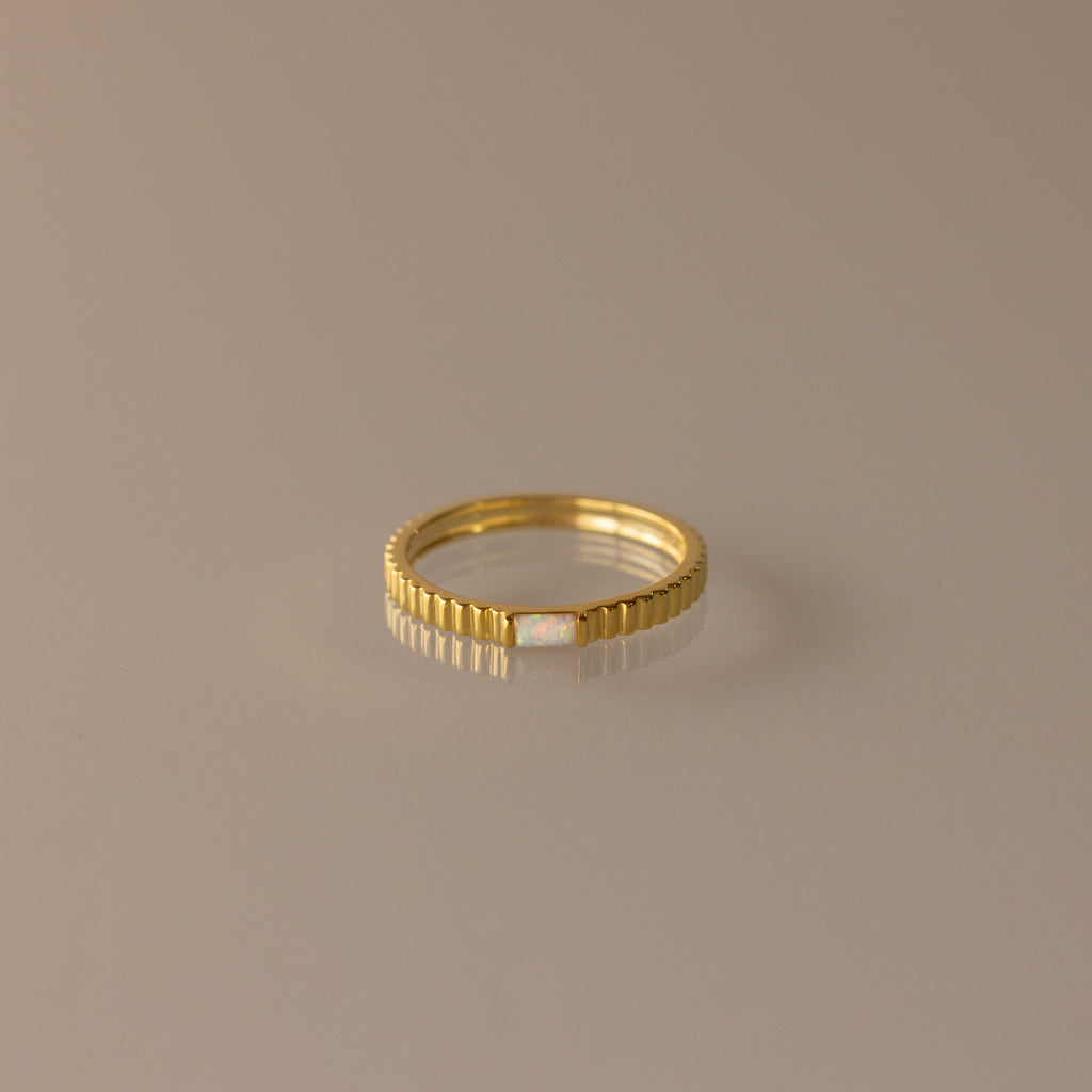 The Dainty Ribbed Opal Ring features a slim gold ribbed band with a small central opal, ideal as a delicate birthstone ring or for stacking, shown on a neutral background.