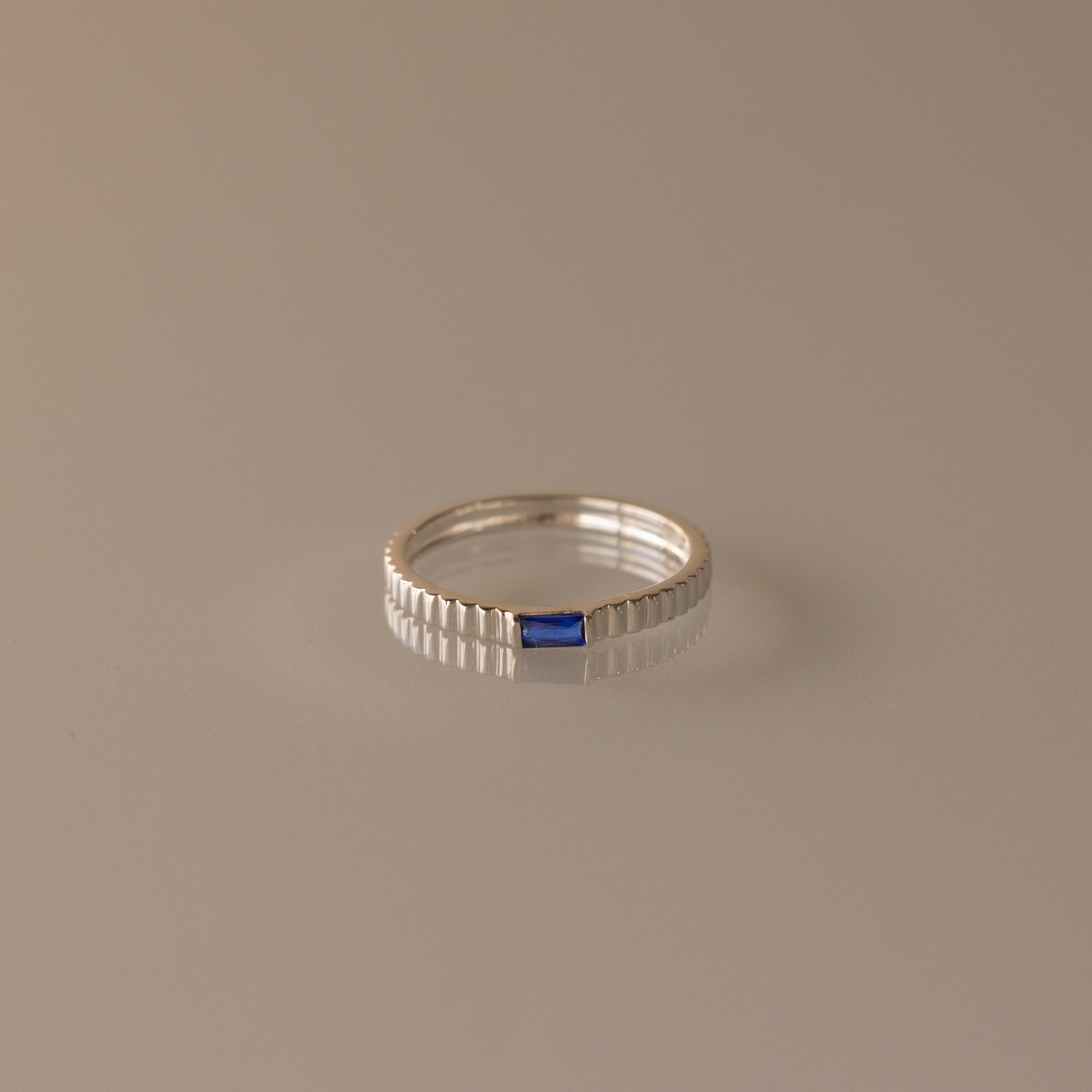 The Dainty Ribbed Sapphire Ring features a ribbed band with a small rectangular blue sapphire centerpiece, ideal for stacking or as a unique birthstone accent.