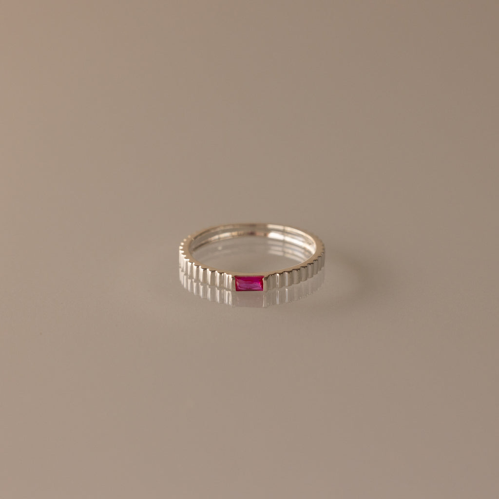 The Dainty Ribbed Ruby Ring is a delicate silver band with ribbed details, featuring a small red ruby at its center, shown against a neutral background.