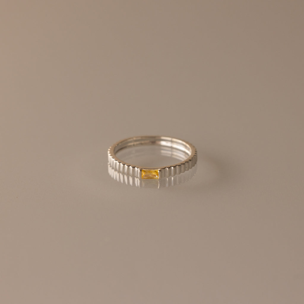 The Dainty Ribbed Topaz Ring is a delicate silver stacking piece with a ridged texture and a small rectangular yellow topaz stone set at the center.