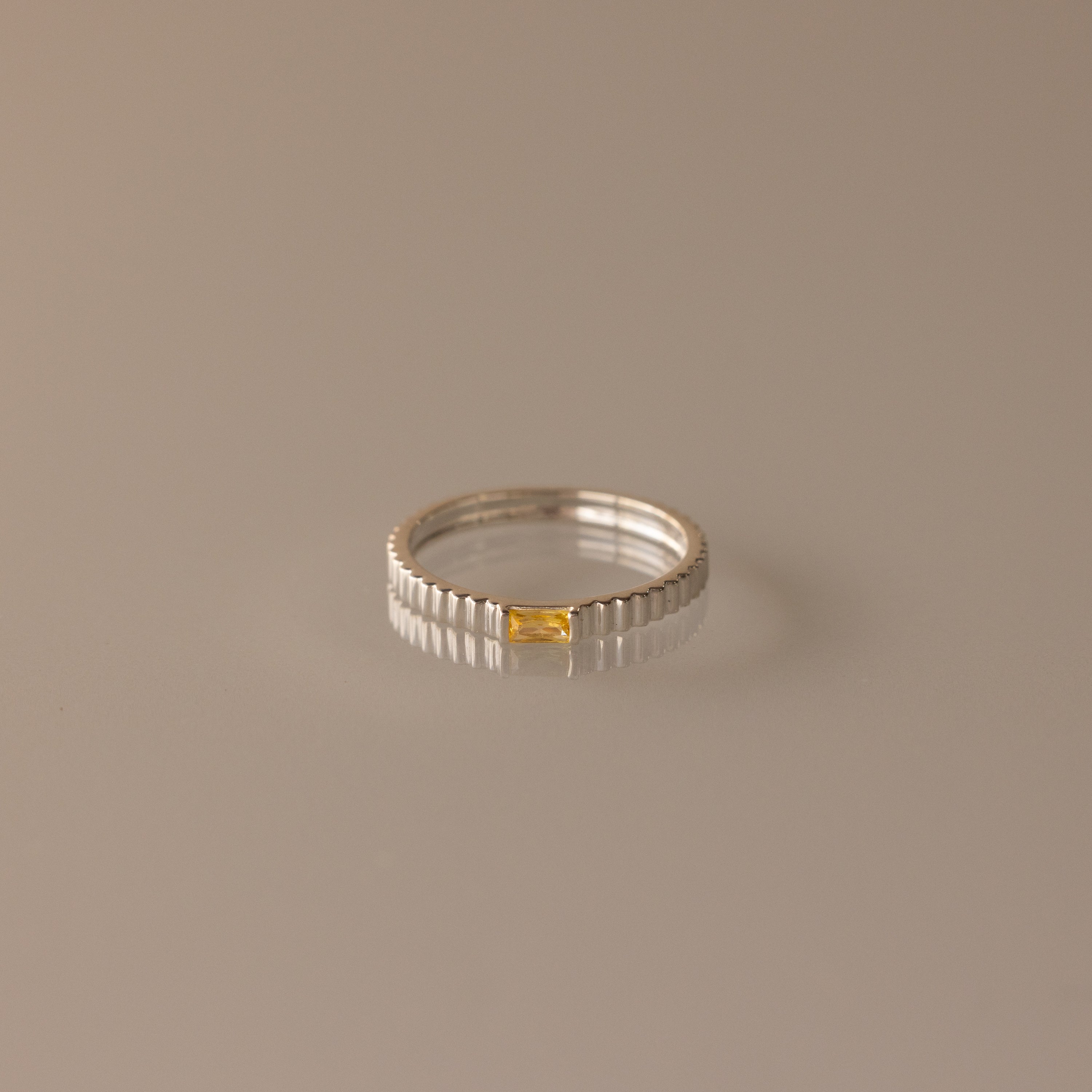 The Dainty Ribbed Topaz Ring is a delicate silver stacking piece with a ridged texture and a small rectangular yellow topaz stone set at the center.