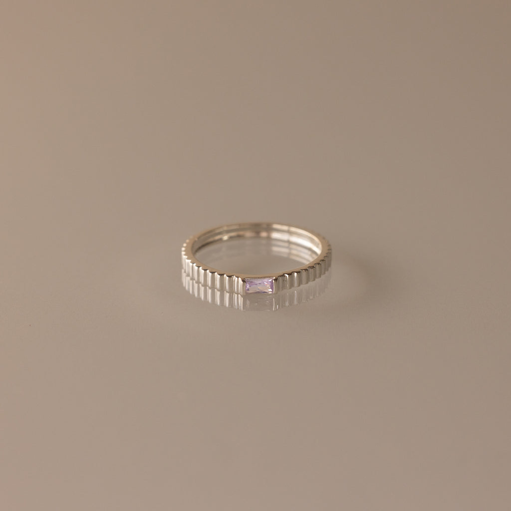 The Dainty Ribbed Alexandrite Ring features a rectangular, light purple gemstone set in silver—ideal as a birthstone ring and shown on a simple beige background.
