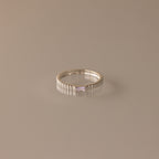 The Dainty Ribbed Alexandrite Ring features a rectangular, light purple gemstone set in silver—ideal as a birthstone ring and shown on a simple beige background.