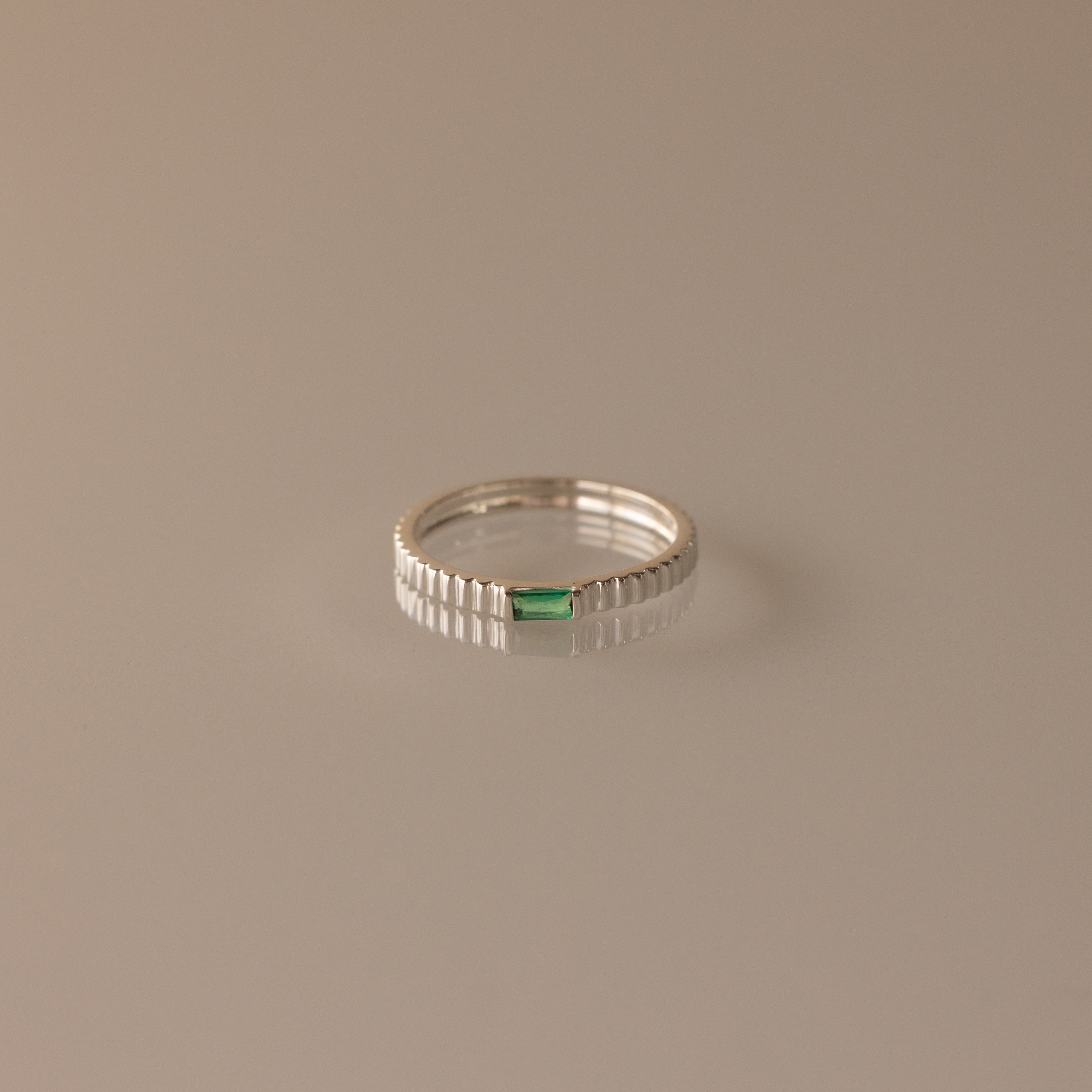 The Dainty Ribbed Emerald Ring showcases a small rectangular green gemstone set at the center of a sleek silver band, beautifully displayed on a beige surface.